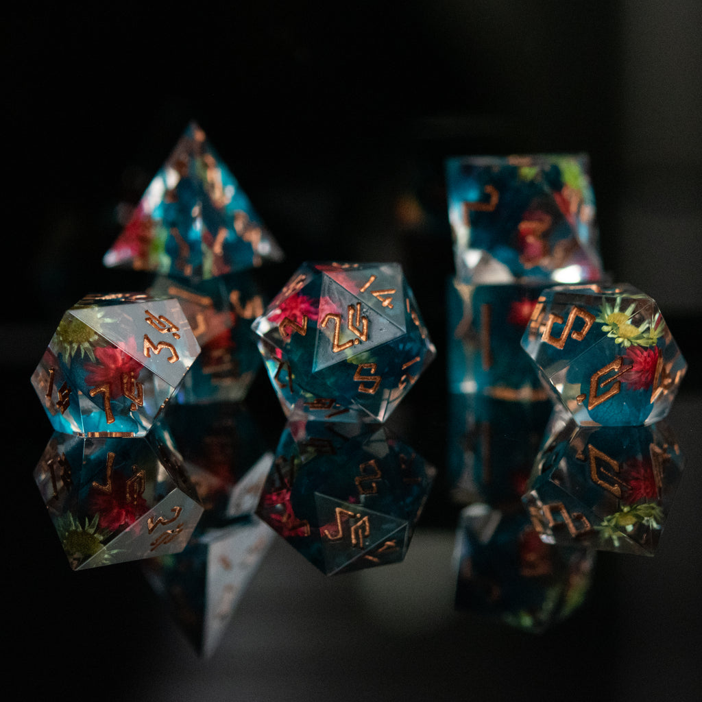 A dice set with blue core and real yellow and pink flowers inside of clear sharp resin featuring gold numbers in a Nordic font