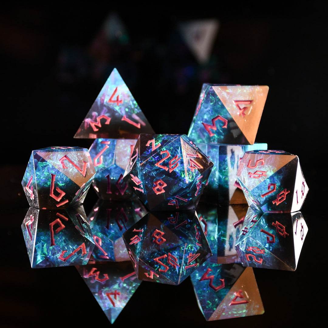 A set of sharp-edged, multicolored resin polyhedral dice with hand-painted pink numbers, with iridescent foil inside reflecting in a mirrored surface.
