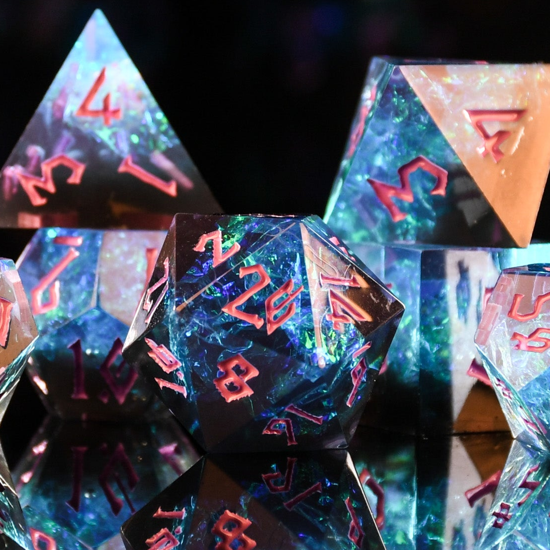 A set of sharp-edged, multicolored resin polyhedral dice with hand-painted pink numbers, with iridescent foil inside reflecting in a mirrored surface.