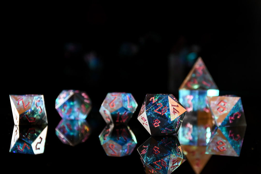 A set of sharp-edged, multicolored resin polyhedral dice with hand-painted pink numbers, with iridescent foil inside reflecting in a mirrored surface.