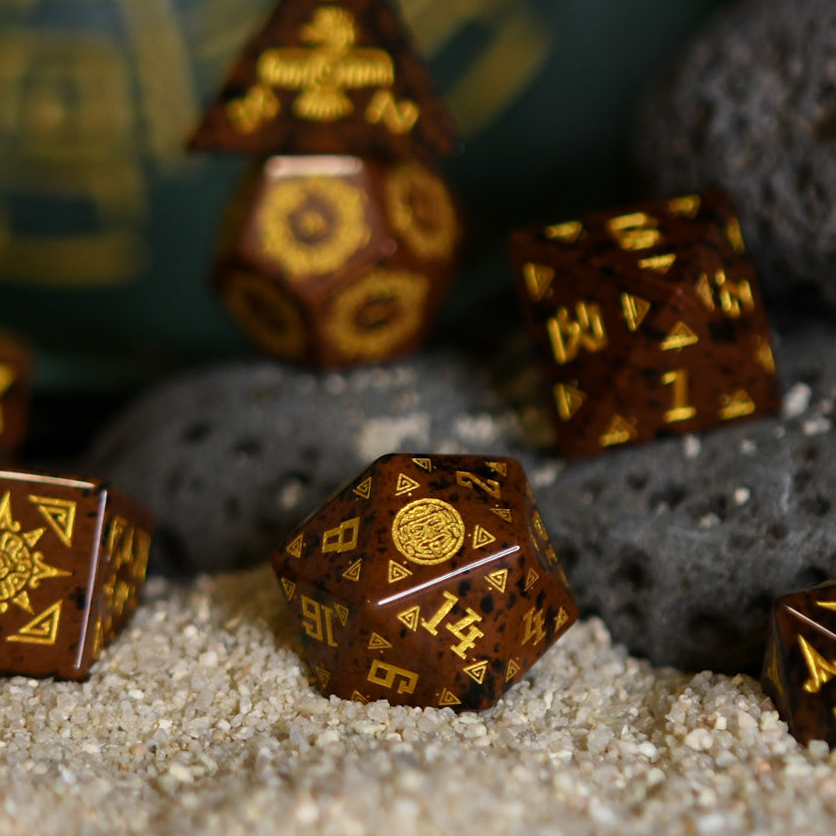 A set of hand-carved, Aztec Mahogany Obsidian Stone Dice with intricate Aztec designs, displayed on a bed of sand.