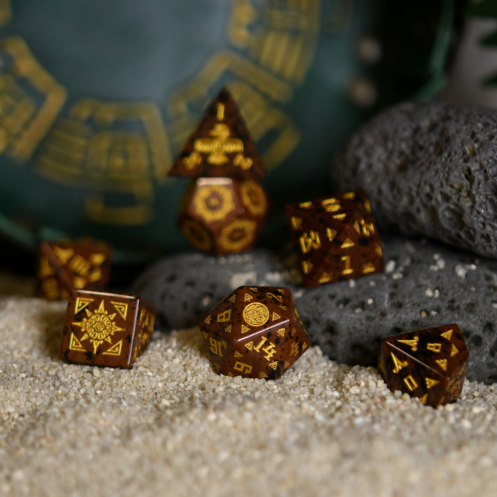 A set of hand-carved, Aztec Mahogany Obsidian Stone Dice with intricate Aztec designs, displayed on a bed of sand.