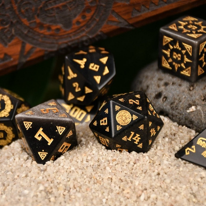 A set of black dice with gold Aztec patterns, placed on sand beside a wooden dice vault box with intricate carvings.