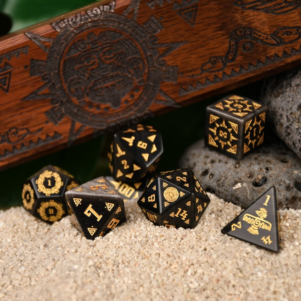 A set of black dice with gold Aztec patterns, placed on sand beside a wooden dice vault box with intricate carvings.