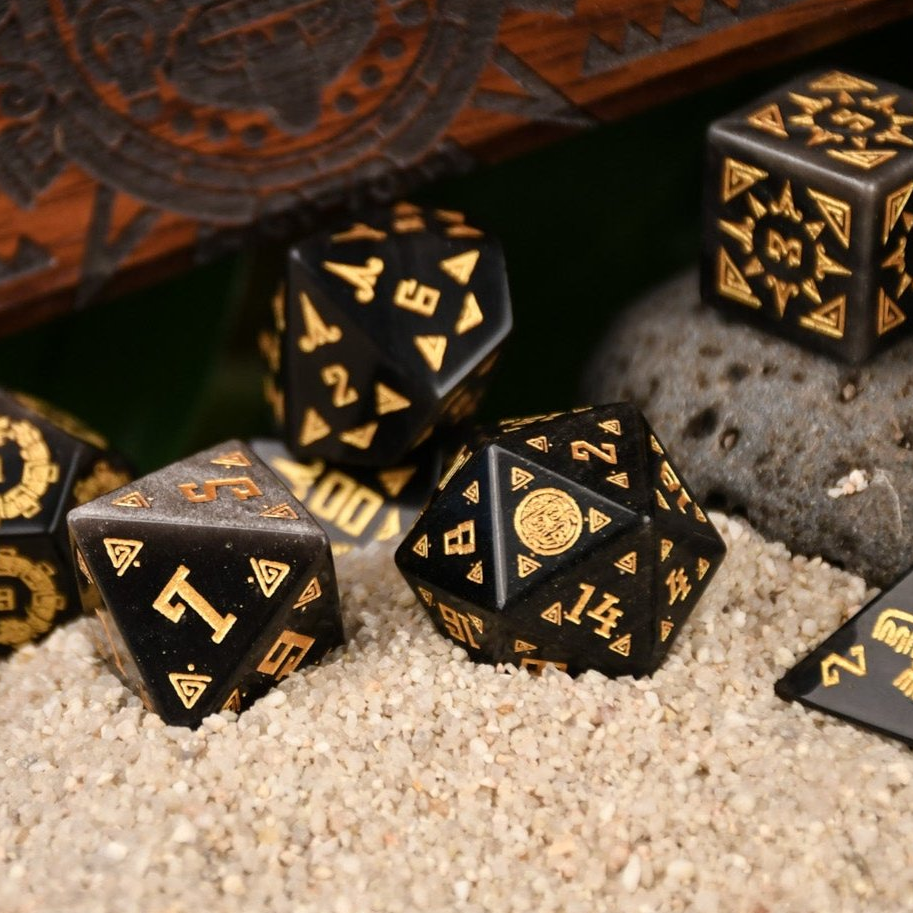 A set of black dice with gold Aztec patterns, placed on sand beside a wooden dice vault box with intricate carvings.