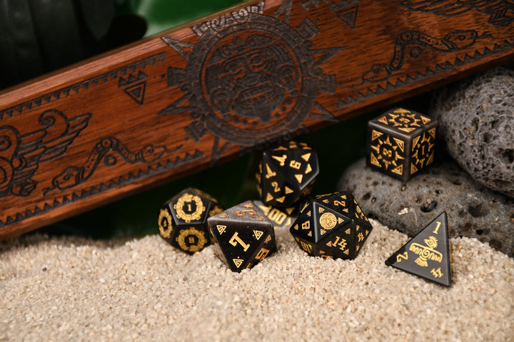 A set of black dice with gold Aztec patterns, placed on sand beside a wooden dice vault box with intricate carvings.
