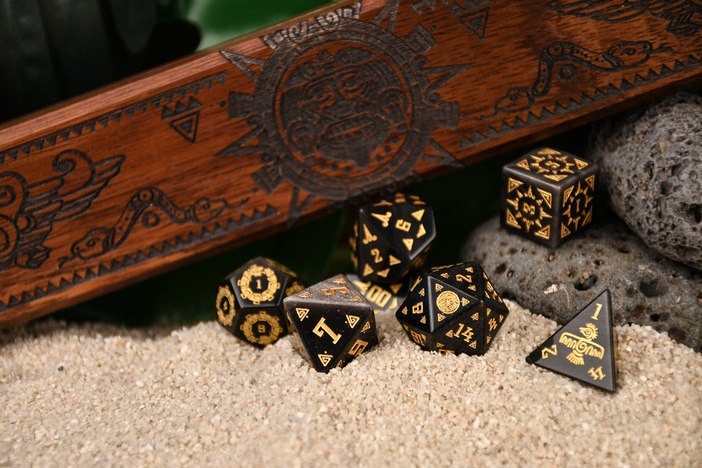 A set of black dice with gold Aztec patterns, placed on sand beside a wooden dice vault box with intricate carvings.