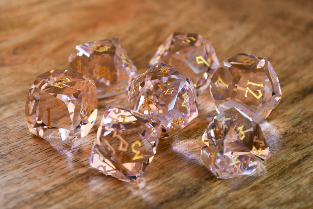 Baby Pink Topaz Multifaceted Glass Dice with gold numbering on wooden surface. 