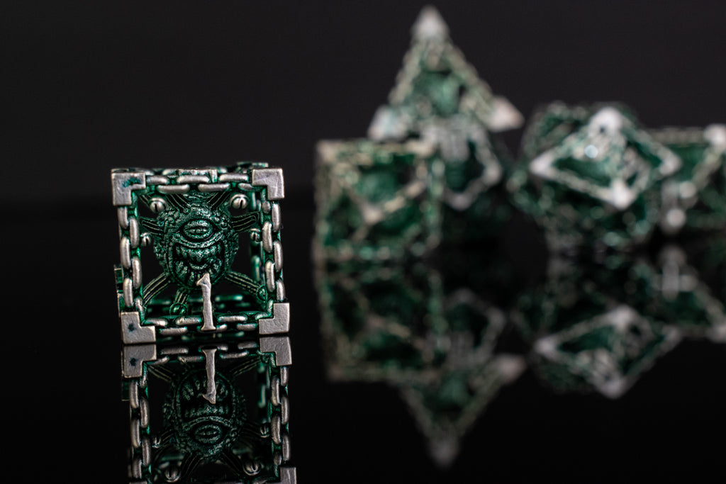 A set of hollow metal dice in green and silver colors with embossed numbers, reflecting on a black surface.