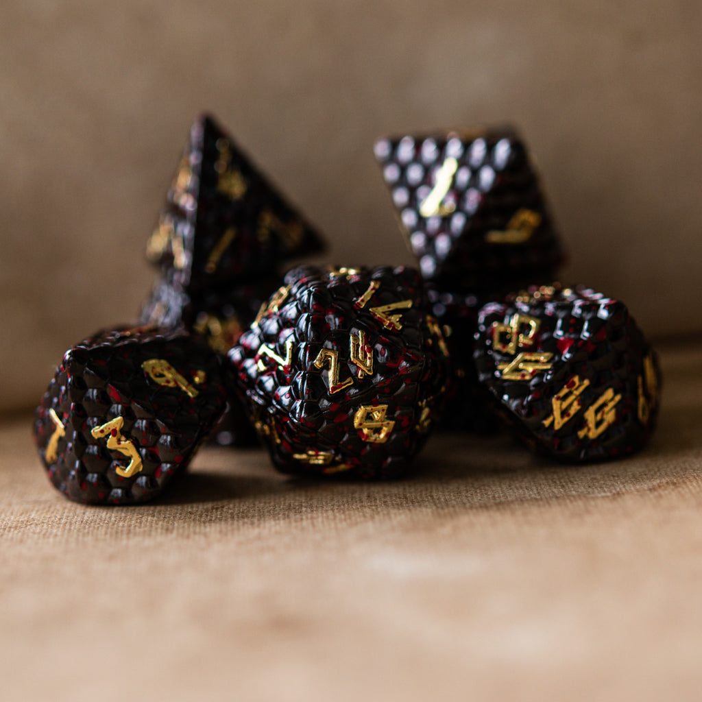 A set of solid metal black dragon scale textured polyhedral dnd dice splattered with blood with gold numbers