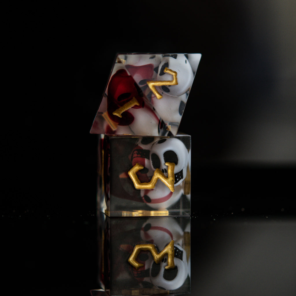 A set of clear sharp-edged resin dice with engraved numbers in gold, with white skulls and red blood inside including various shapes such as a d4, d6, d8, d10, d12, and d20 for tabletop and board games