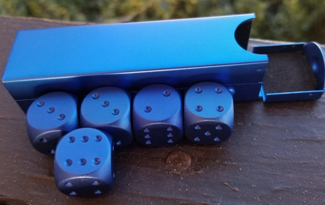 A set of blue aluminum D6 dice with engraved numbers and pips, displayed in front of a foam-lined case.