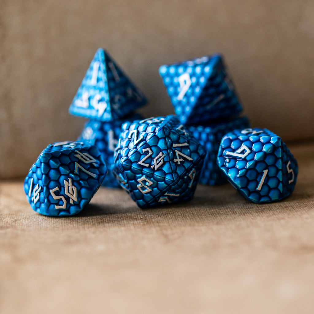 A set of blue dragon scale texture metal dice with embossed numbers, including various shapes such as a d4, d6, d8, d10, d12, and d20, displayed on a textured surface.