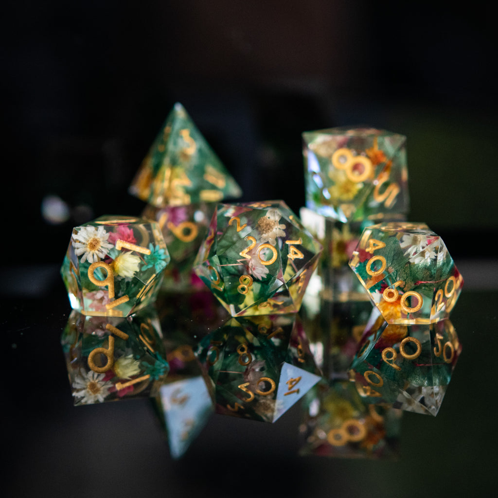 A set of sharp-edged, hand-painted resin dnd dice with floral and botanical designs, featuring gold, green, pink, and yellow colors for tabletop gaming and board games