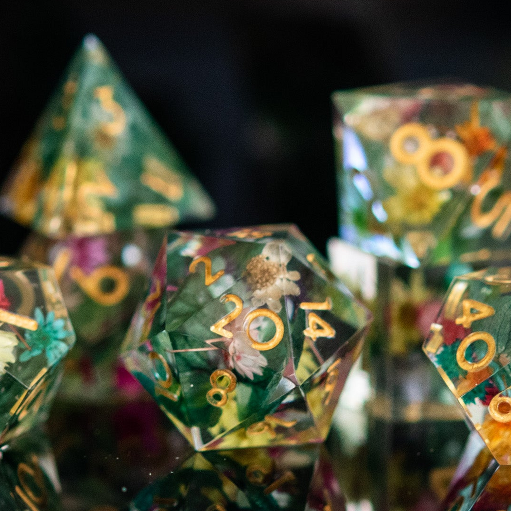 A set of sharp-edged, hand-painted resin dnd dice with floral and botanical designs, featuring gold, green, pink, and yellow colors for tabletop gaming and board games