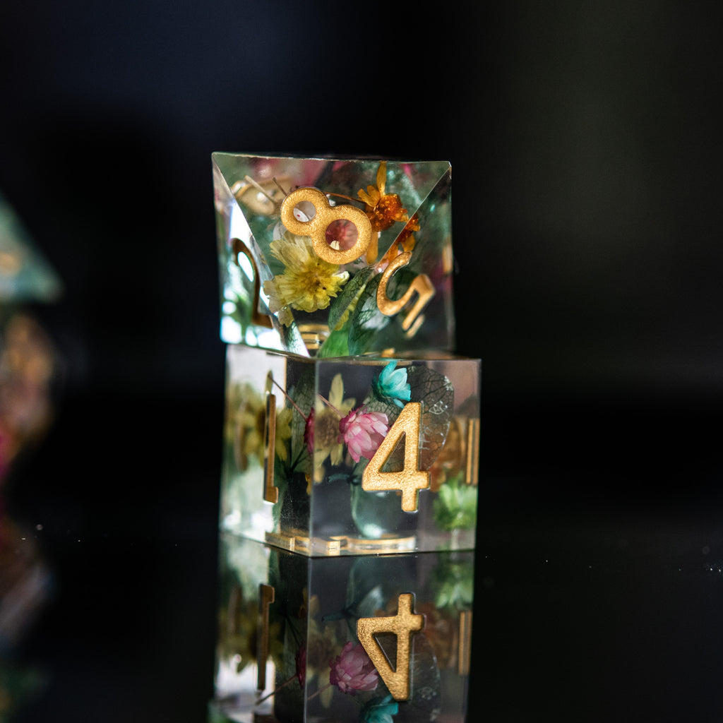 A set of sharp-edged, hand-painted resin dnd dice with floral and botanical designs, featuring gold, green, pink, and yellow colors for tabletop gaming and board games