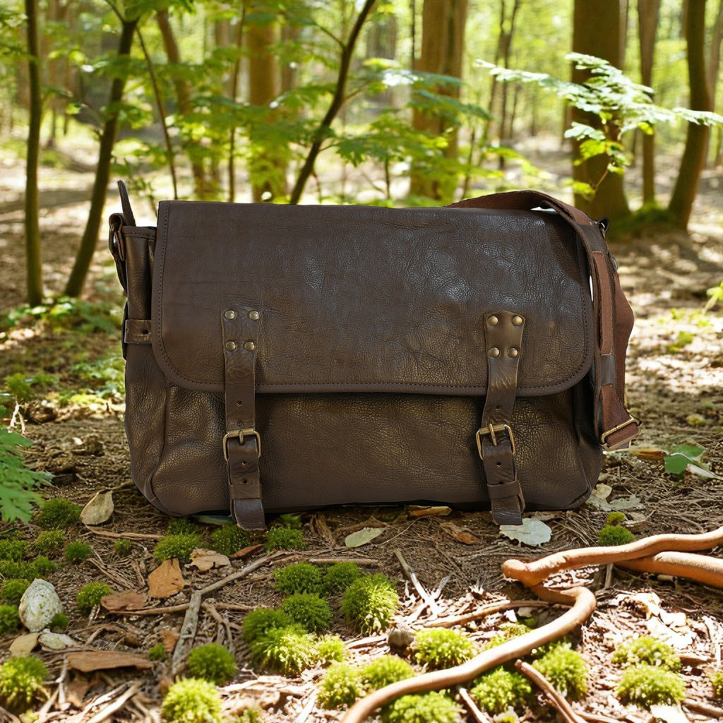 Brown leather modern rugged messenger bag on a forest floor with trees and greenery in the background