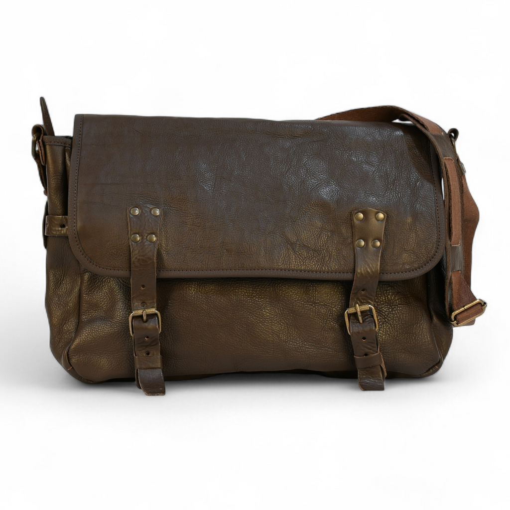 Brown leather modern rugged messenger bag on a forest floor with trees and greenery in the background