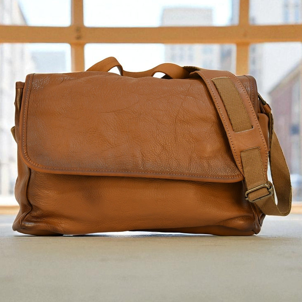 Brown leather modern rugged messenger bag on a surface with a window in the background