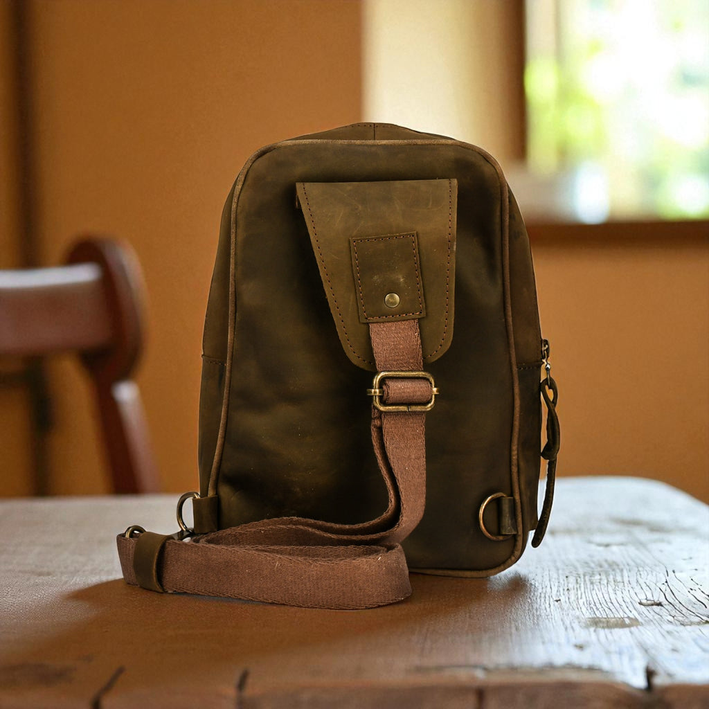 olive leather sling bag on a wooden surface with books and plants in the background