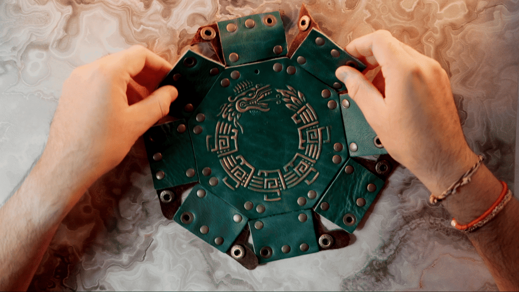 An octagonal Celtic knot leather dice tray with snap buttons and a gold-colored knot design in the center, displayed on a wooden surface.
