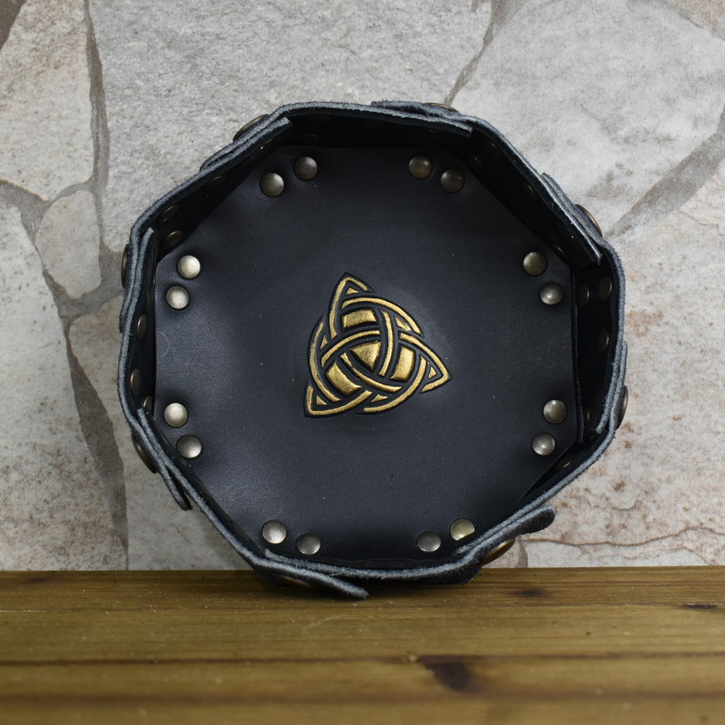 An octagonal Celtic knot leather dice tray with snap buttons and a gold-colored knot design in the center, displayed on a wooden surface.