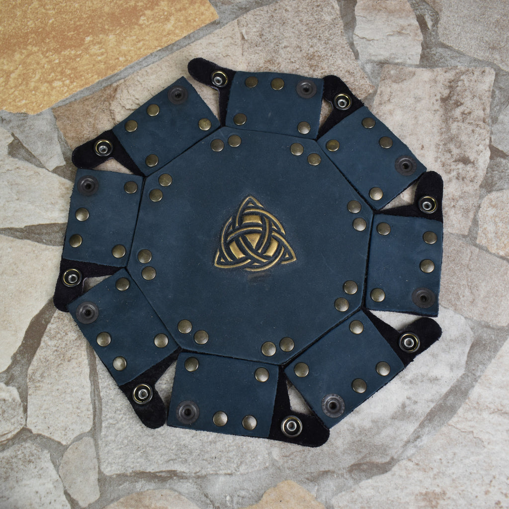 An octagonal Celtic knot leather dice tray with snap buttons and a gold-colored knot design in the center, displayed on a wooden surface.