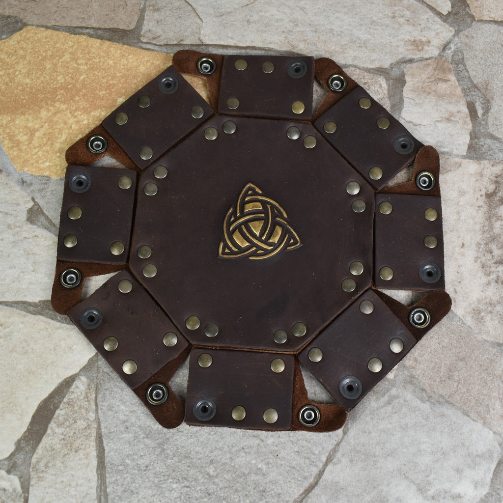 An octagonal Celtic knot leather dice tray with snap buttons and a gold-colored knot design in the center, displayed on a wooden surface.