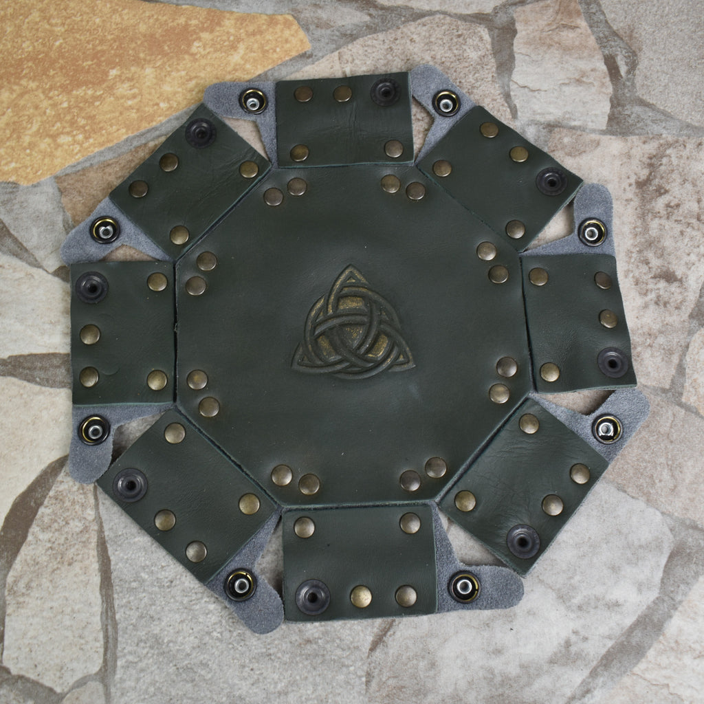 An octagonal Celtic knot leather dice tray with snap buttons and a gold-colored knot design in the center, displayed on a wooden surface.