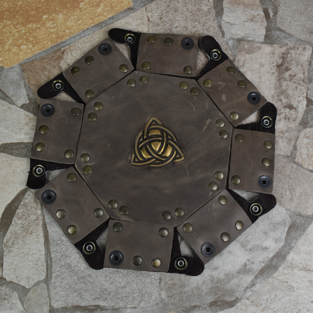 An octagonal Celtic knot leather dice tray with snap buttons and a gold-colored knot design in the center, displayed on a wooden surface.