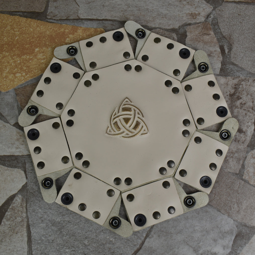 An octagonal Celtic knot leather dice tray with snap buttons and a gold-colored knot design in the center, displayed on a wooden surface.