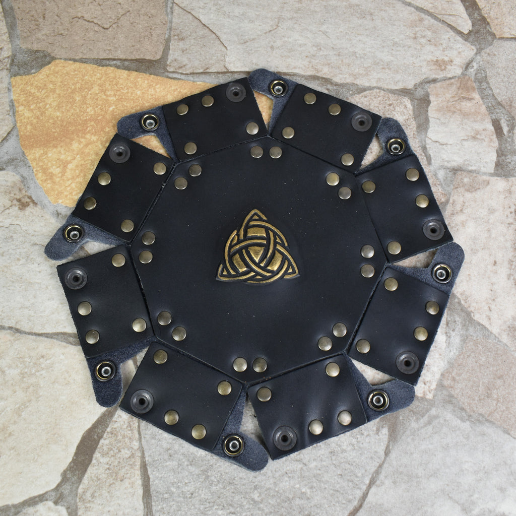 An octagonal Celtic knot leather dice tray with snap buttons and a gold-colored knot design in the center, displayed on a wooden surface.