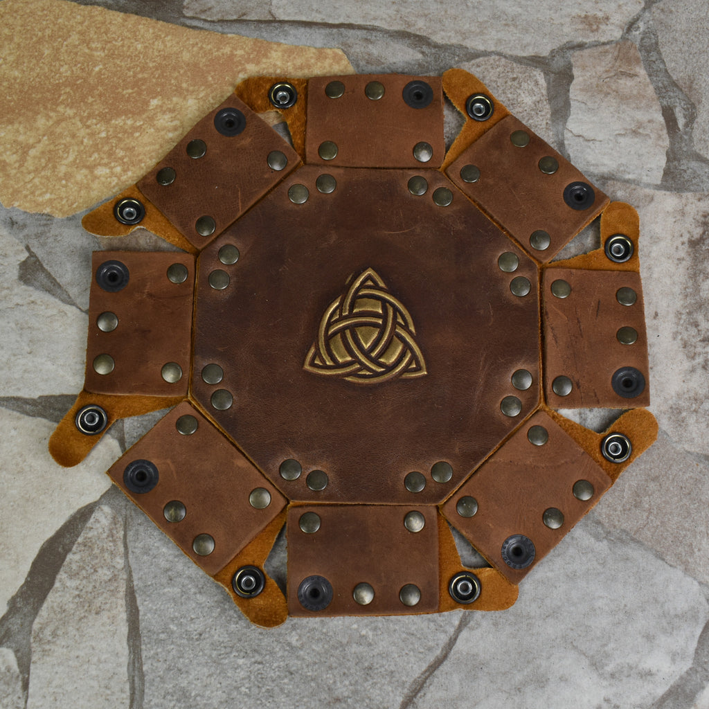 An octagonal Celtic knot leather dice tray with snap buttons and a gold-colored knot design in the center, displayed on a wooden surface.