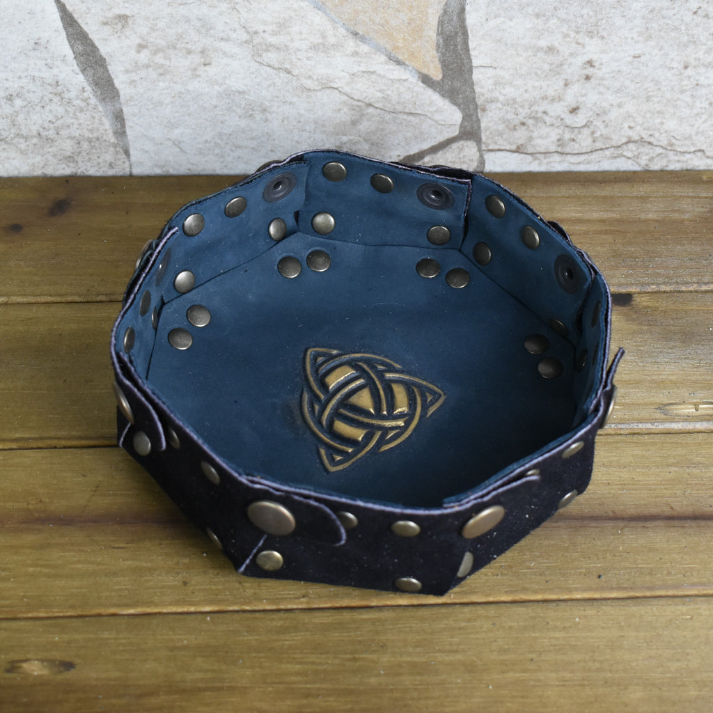 An octagonal Celtic knot leather dice tray with snap buttons and a gold-colored knot design in the center, displayed on a wooden surface.