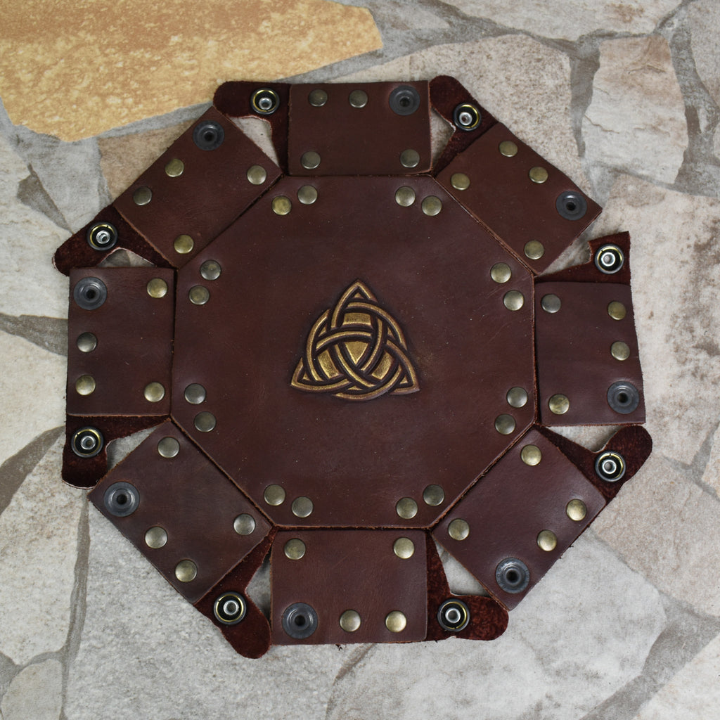 An octagonal Celtic knot leather dice tray with snap buttons and a gold-colored knot design in the center, displayed on a wooden surface.