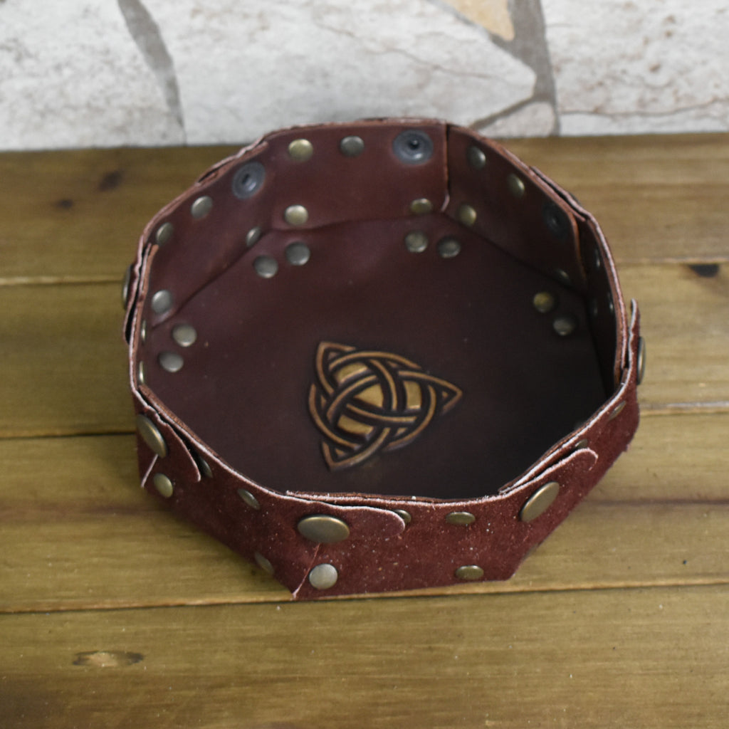 An octagonal Celtic knot leather dice tray with snap buttons and a gold-colored knot design in the center, displayed on a wooden surface.