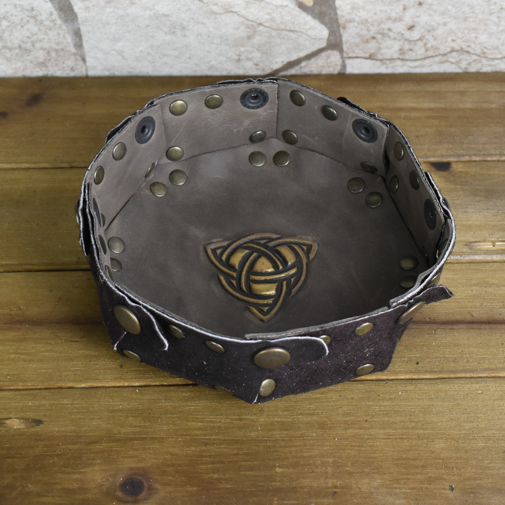An octagonal Celtic knot leather dice tray with snap buttons and a gold-colored knot design in the center, displayed on a wooden surface.