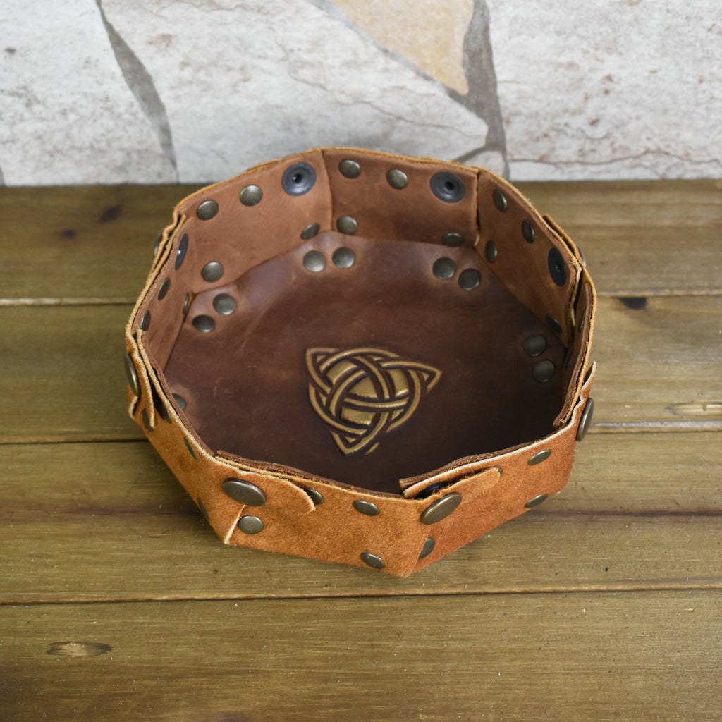 An octagonal Celtic knot leather dice tray with snap buttons and a gold-colored knot design in the center, displayed on a wooden surface.