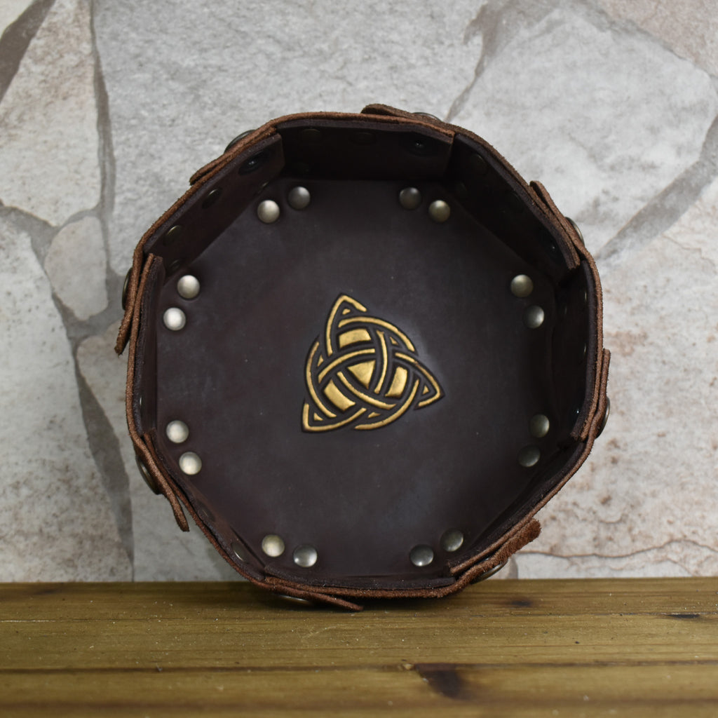 An octagonal Celtic knot leather dice tray with snap buttons and a gold-colored knot design in the center, displayed on a wooden surface.