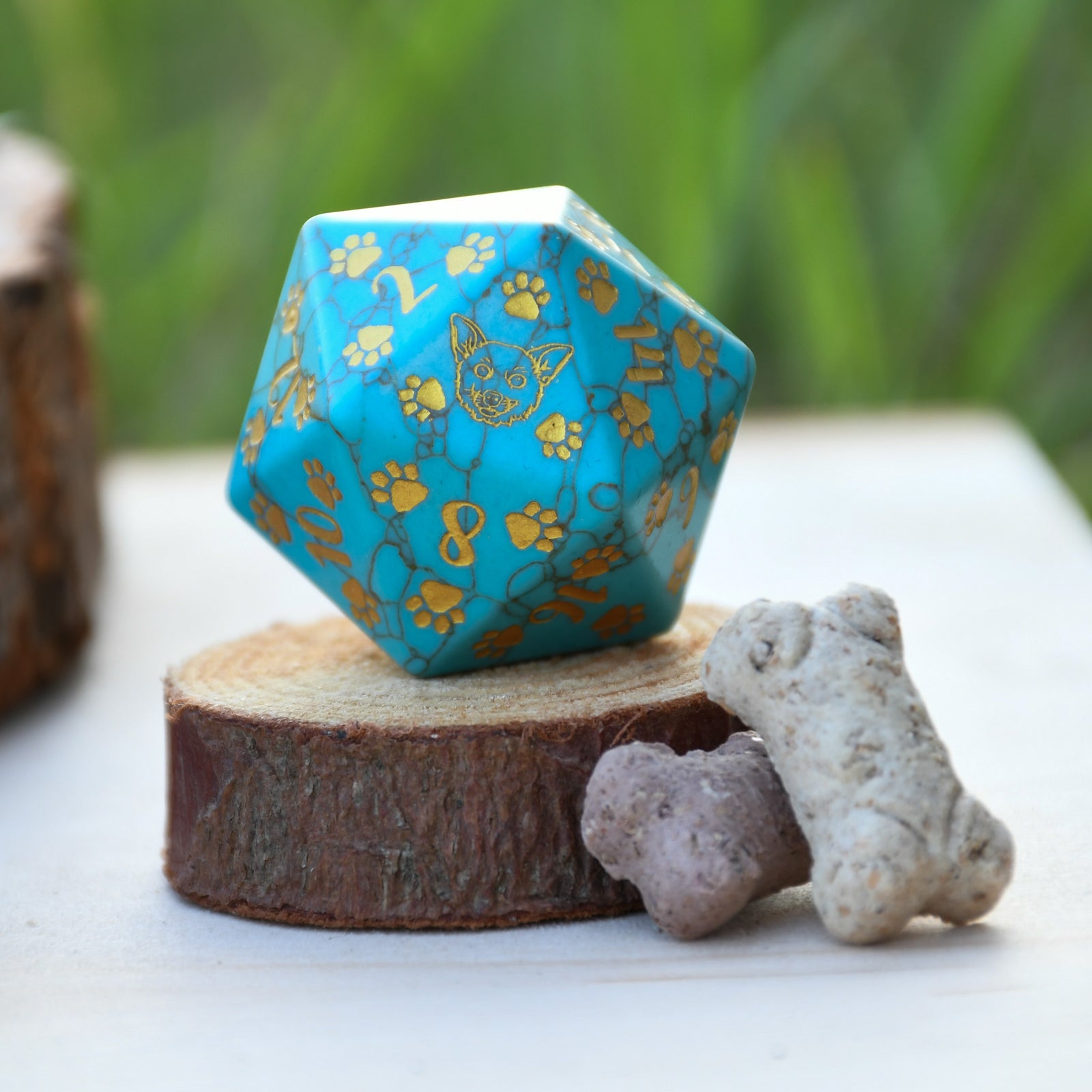 A hand-carved gemstone D20 dice with green turquoise coloring and decorative elements in the form of a puppy paw and bone on one of the faces, placed on a wooden surface next to a piece of driftwood.