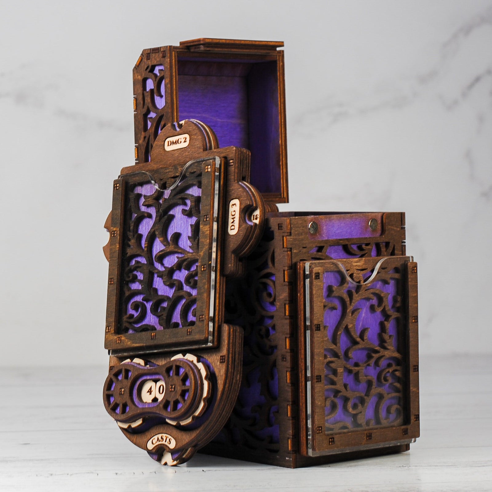 A wooden deck box with intricate cut-out patterns, featuring a purple interior, accompanied by a command plate and game tokens.