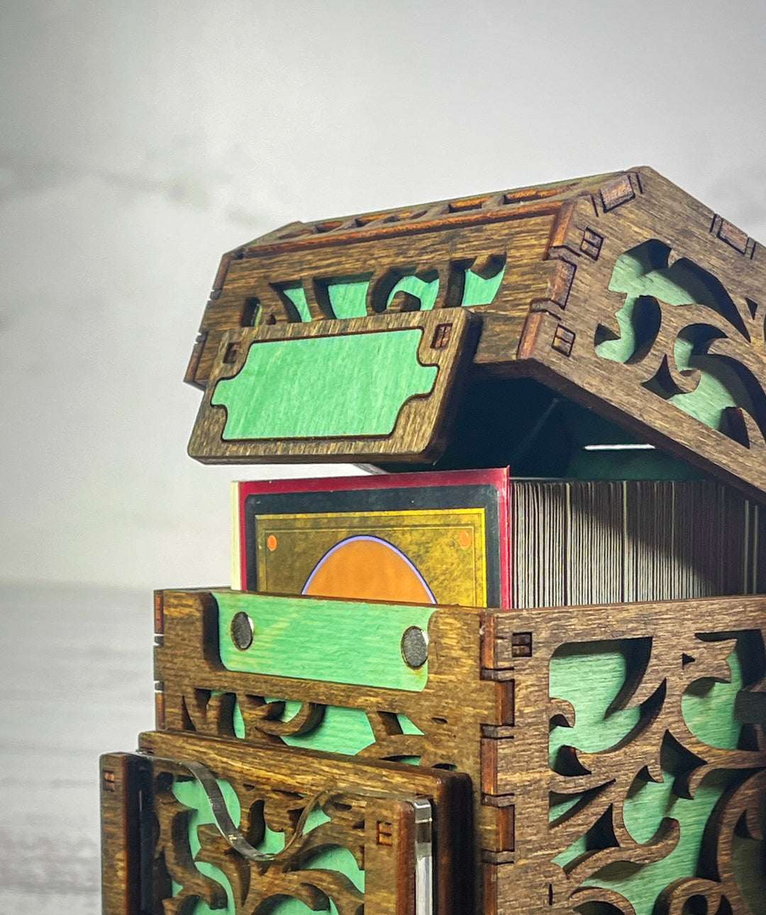 A wooden deck box with a intricate carved design, featuring a removable magnetic plate on the front for holding cards from the game &