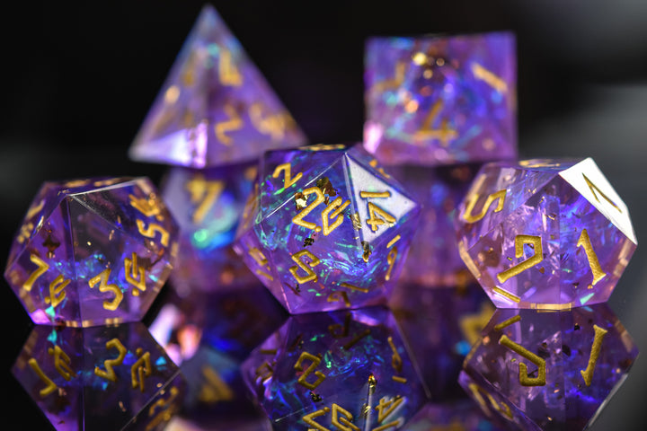 A set of sharp-edged, iridescent purple resin dice with hand-painted blue numbers, reflecting light and displaying a shiny surface.