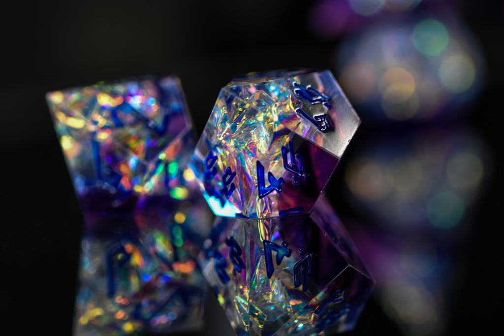 A set of sharp-edged, iridescent purple resin dice with hand-painted blue numbers, reflecting light and displaying a shiny surface.