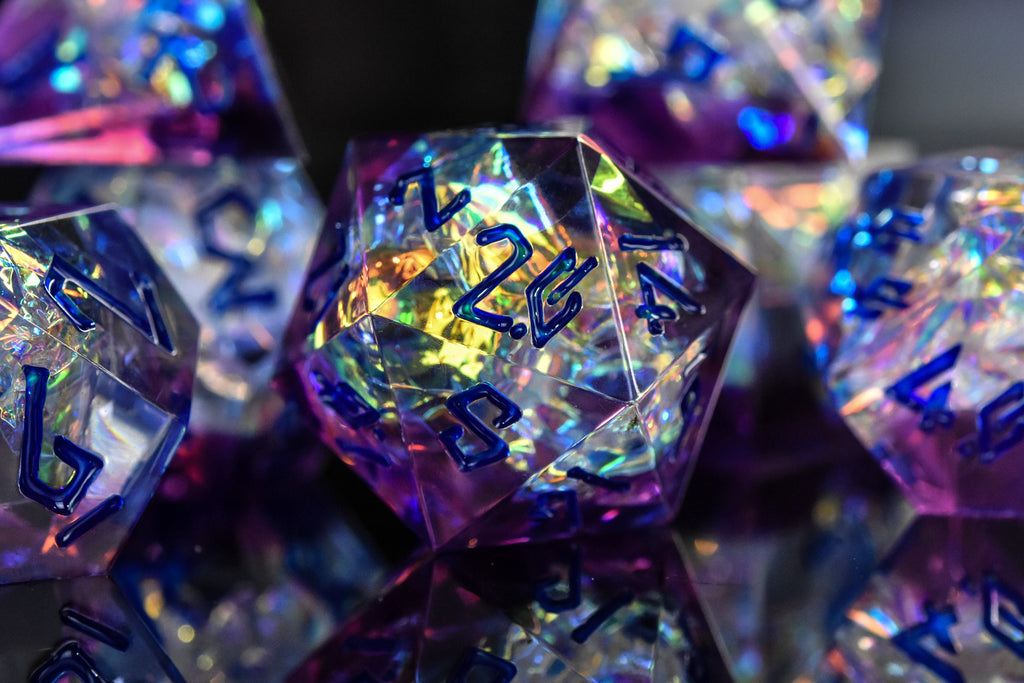 A set of sharp-edged, iridescent purple resin dice with hand-painted blue numbers, reflecting light and displaying a shiny surface.