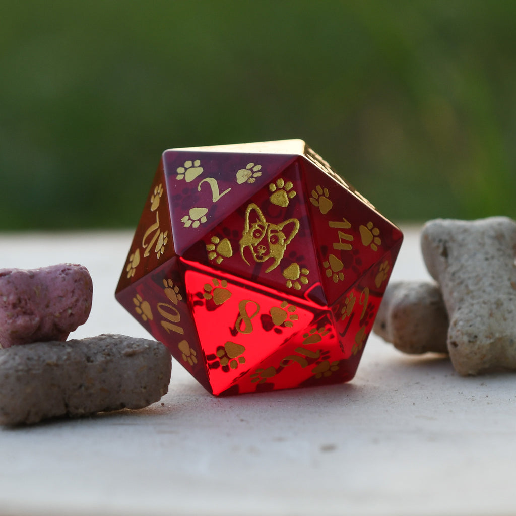A red glass D20 dice with gold paw prints and dog designs hand painted on it, displayed on a white surface with rocks in the background.