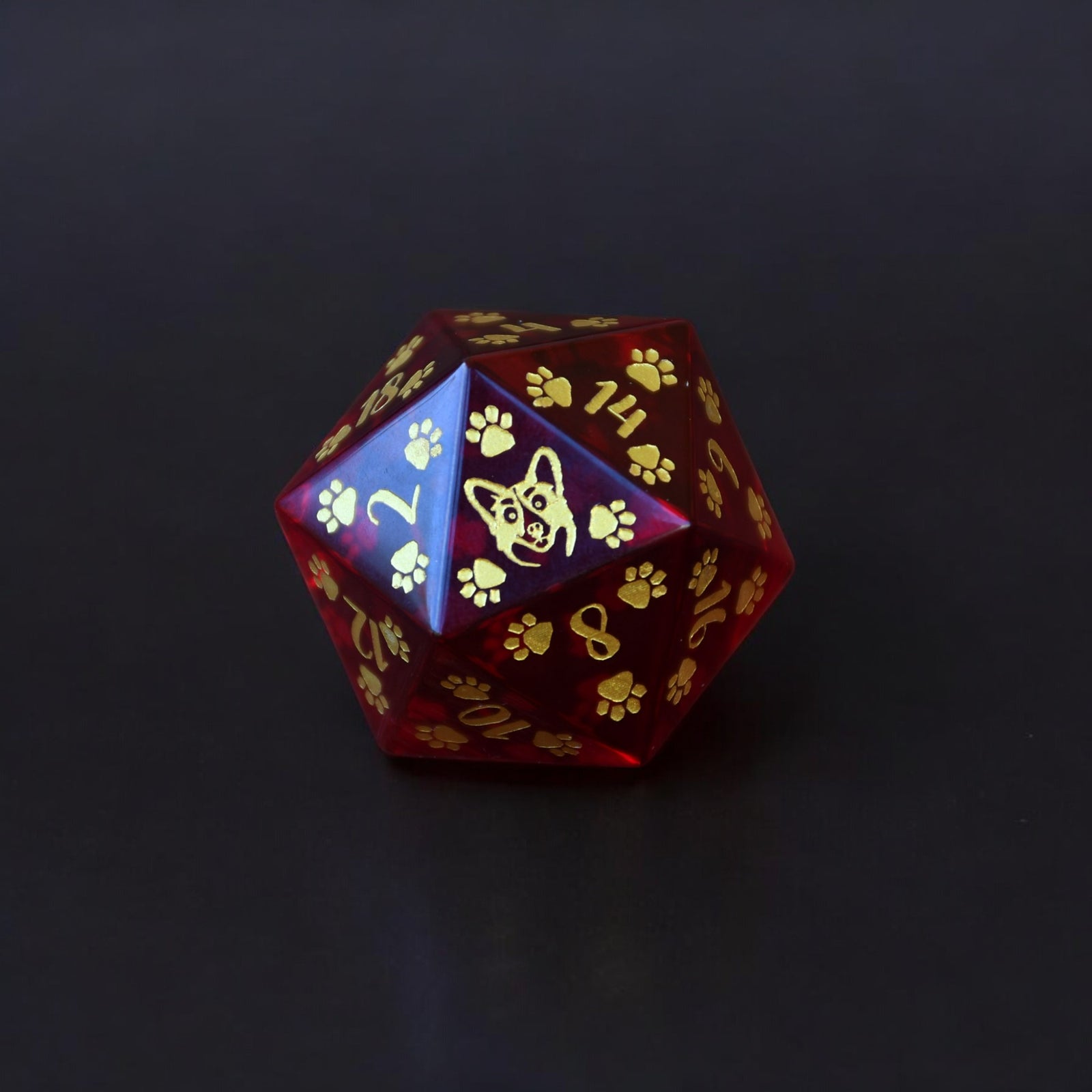 A red glass D20 dice with gold paw prints and dog designs hand painted on it, displayed on a white surface with rocks in the background.