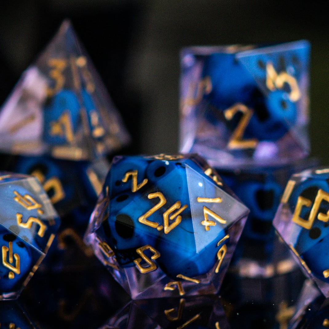 A set of sharp-edged resin dice in blue and lavender with engraved numbers, with blue skulls inside including various shapes such as a d4, d6, d8, d10, d12, and d20.