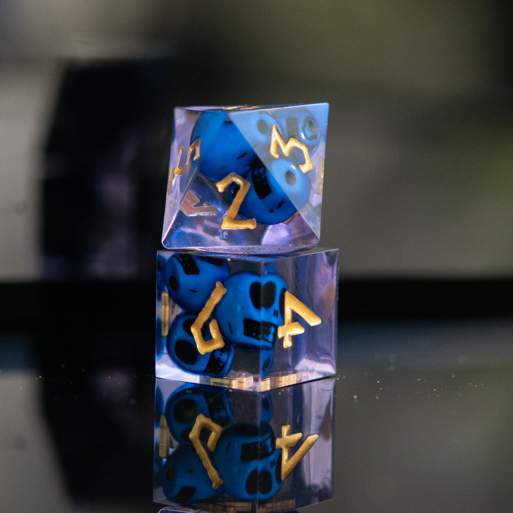 Stacked sharp-edged resin dice in blue and lavender with engraved numbers, with blue skulls inside including various shapes such as a d4, d6, d8, d10, d12, and d20.
