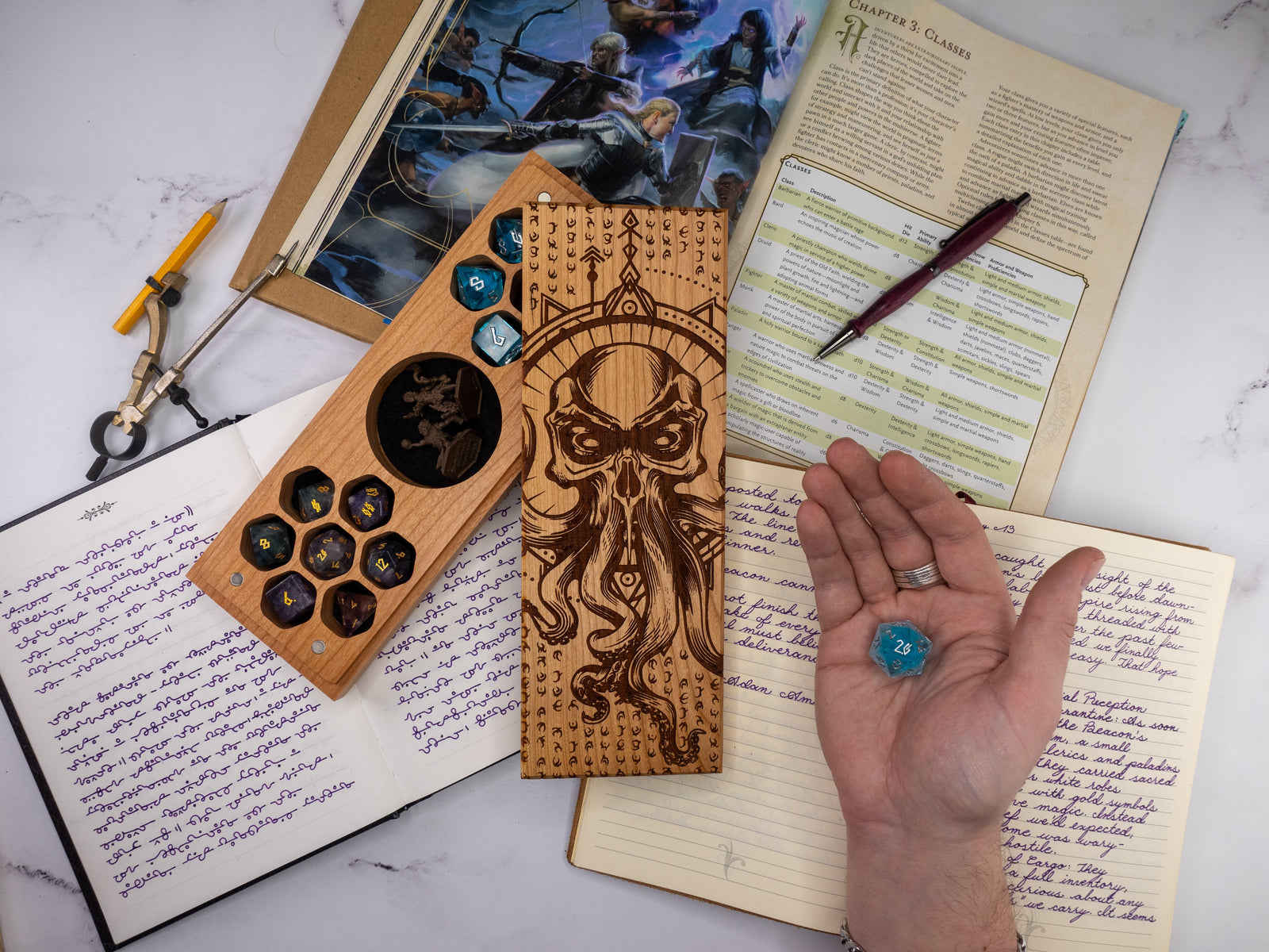 Wooden dice tray with dice and a hand holding a blue die on top of books and papers.
